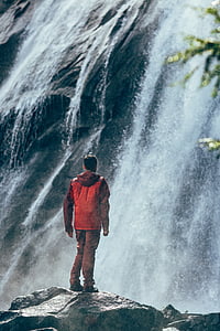 man wearing red hooded windbreaker jacket standing on gray rock while watching waterfalls at daytime thumbnail
