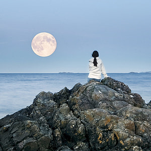 woman wearing white hoodie on top of rock facing towards the full moon thumbnail