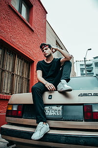 man sitting on brown car's trunk parked near red concrete building at daytime thumbnail