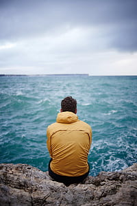 man in yellow hooded jacket sitting on cliff in front of sea thumbnail
