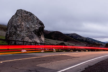 timelapse photography of vehicle passing on road near giant rock under cloudy sky thumbnail