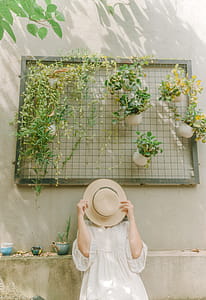 Woman Wearing White Dress Leaning on White Wall Covering Her Face With Brown Hat thumbnail