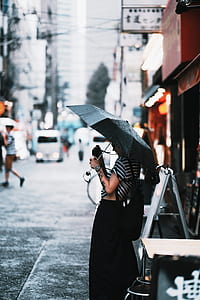 woman holding umbrella and point-and-shoot camera while standing beside table and signage in street during daytime thumbnail