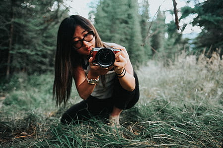 woman in grey shirt and black pants holding grey and black camera sitting on green grass selective-focus photography thumbnail