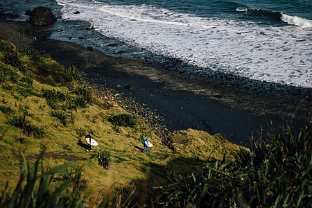 aerial photography of two person walking down hill towards beach shore while carrying surfboards thumbnail