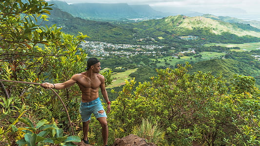 man topless wearing blue denim distress bermuda shorts holding brown rope while watching aerial view on mountain and ocean at daytime thumbnail