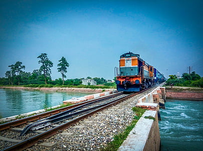 orange and black train passing on rail road between body of water under blue sky thumbnail