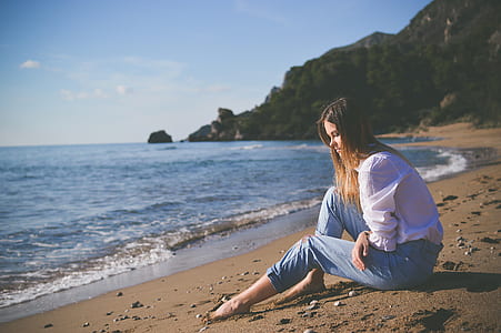 woman sitting on shore beside large body of water and mountains during daytime thumbnail