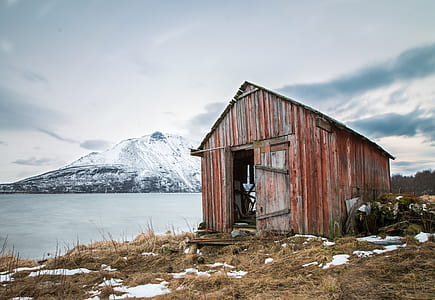 brown barn beside large body of water near mountain covered with snow under white and blue cloudy skies thumbnail