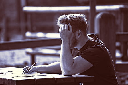 man sitting at table with left hand near face and right hand holding paper and pen thumbnail