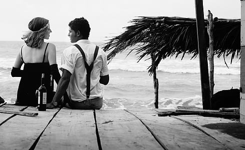 grayscale photo of couple sitting on wooden surface near seashore at daytime thumbnail