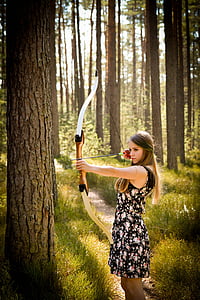 woman in black floral mini dress holding long bow standing beside tree thumbnail