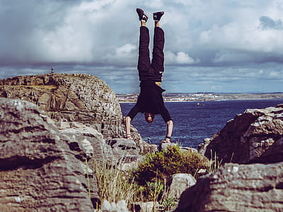Man Wearing Black Dress Shirt and Burgundy Pants Hand Standing on Rock Formations Across Sea thumbnail