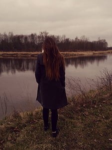 back view of woman wearing black coat and bottoms in front of body of water thumbnail