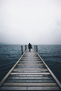 man in black suit walks on brown wooden dock in middle of sea under cloudy sky during daytime thumbnail