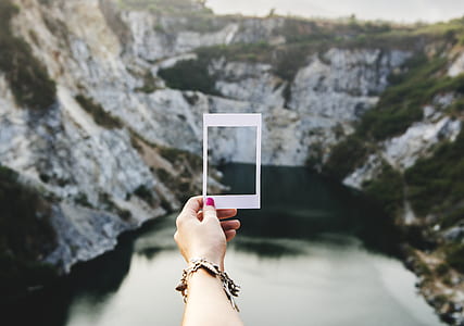 person holding white rectangular frame in front of grey calm body of water and rock formations during daytime thumbnail