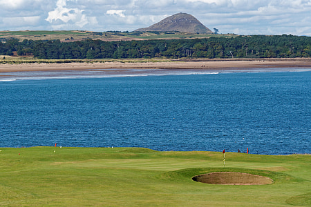 golf field near body of water under white clouds and blue sky thumbnail