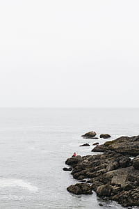 person sitting on gray rock beside body of water thumbnail
