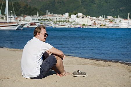 Photo of Man in White Shirt and Blue Short Sitting on Seashore thumbnail