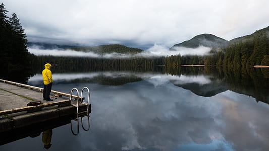 man standing on corner of dock facing body of water with trees and mountain in background thumbnail