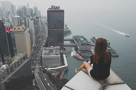 woman sitting on building while looking at cityscape near body of water thumbnail