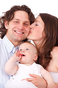 Brown Haired Woman Kissing Man in Blue White Dress Shirt Holding Baby in White Dress thumbnail