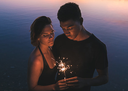 man and woman in black top holding together sparkler light near body of wtaer thumbnail