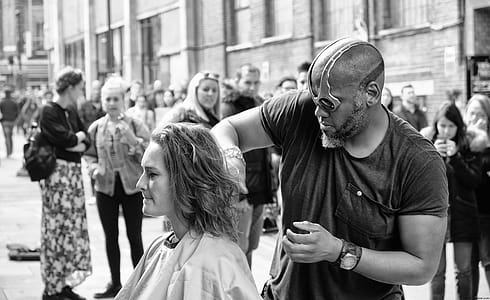 Grayscale Photography of Man Cutting Hair of Woman Surrounded With People thumbnail