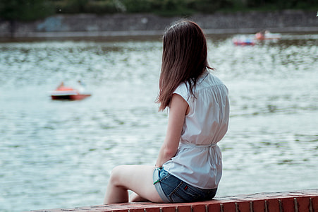 woman wearing white sleeveless dress sitting on brown brick near body of water during daytime thumbnail