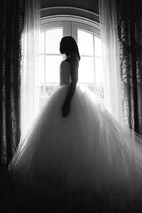 grayscale photography of woman wearing strapless ball gown wedding dress standing and facing in front of arch window in between floral curtains thumbnail