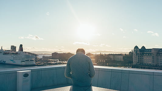 man wearing quilted gray jacket sitting on building top across body of water with docked cruise ship beside buildings under blue sky thumbnail