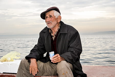 man wearing leather jacket holding plastic cup sitting on concrete by the sea thumbnail