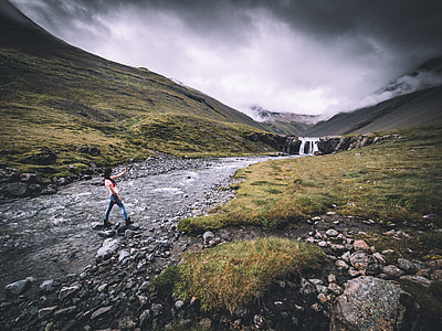woman wearing pink shirt and blue denim jeans standing on river between green grass field under gray and white cloudy sky thumbnail