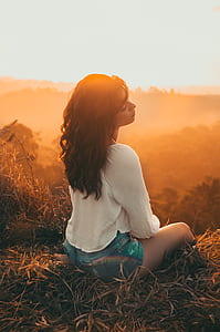 Woman in White Long-sleeved Shirt With Blue Short Shorts Sitting on Brown Grass Against Golden Hour Light thumbnail