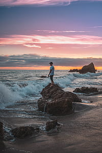 man standing on brown rock formation beside seashore during sunset thumbnail