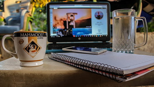 White Smartphone on Spiral Notebook Between Ceramic Mug and Glass Beer Mug Near Black Computer Monitor Showing Hour Glass thumbnail