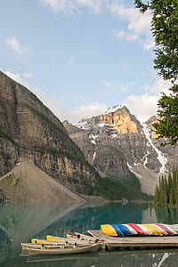 assorted-color canoe lot beside body of water near rocky mountain under blue sky thumbnail