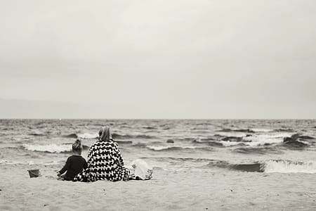 Woman And Child Sitting On Sand Near Body Of Water thumbnail
