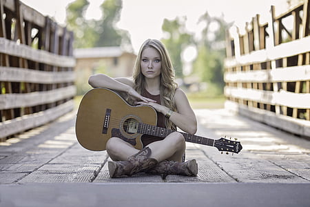 selective focus photography of woman wearing sleeveless top holding cutaway acoustic guitar sitting on bridge thumbnail