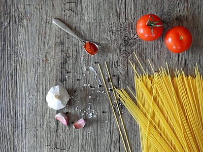 top angle photo of uncooked pasta beside two tomatoes, garlic, and spoon of red powder above wooden surface thumbnail