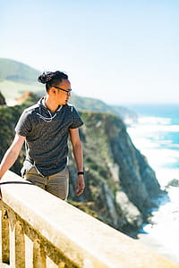 man holding balustrade near green mountain with sea below thumbnail