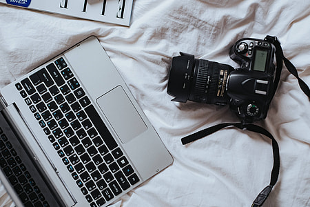 Silver laptop, a camera, magazines and other items on white bed sheets thumbnail
