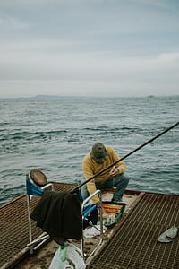 man wearing yellow sweatshirt and blue denim jeans outfit sitting beside body of water under white sky at daytime thumbnail