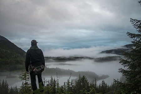 man with black hooded jacket standing beside pine tree watching mountain view during daytime thumbnail