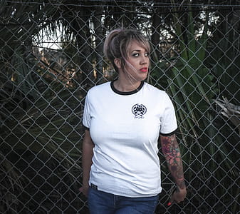 Woman Wearing White Crew-neck T-shirt With Black Trim and Blue Denim Bottoms Beside Wire Fence thumbnail