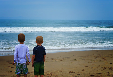 two boys standing on brown sand in front body of water under blue sky thumbnail
