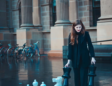 woman standing while holding two black chess pieces in front of brown concrete building during daytime thumbnail