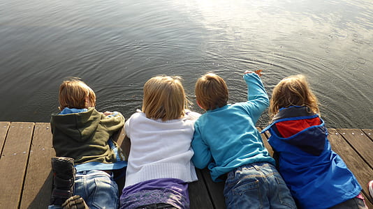 four children lying on brown deck while staring at body of water thumbnail