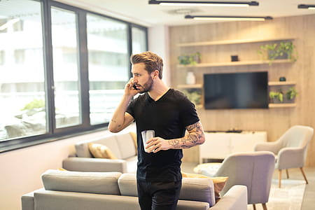 A man in a black tshirt talks on a cell phone while standing with a drink in a living room. thumbnail