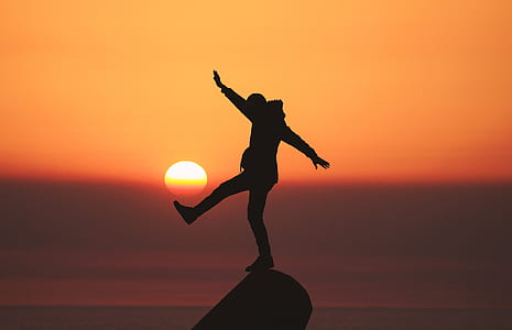 silhouette of person standing with one foot on rock surrounded by water during golden hour thumbnail